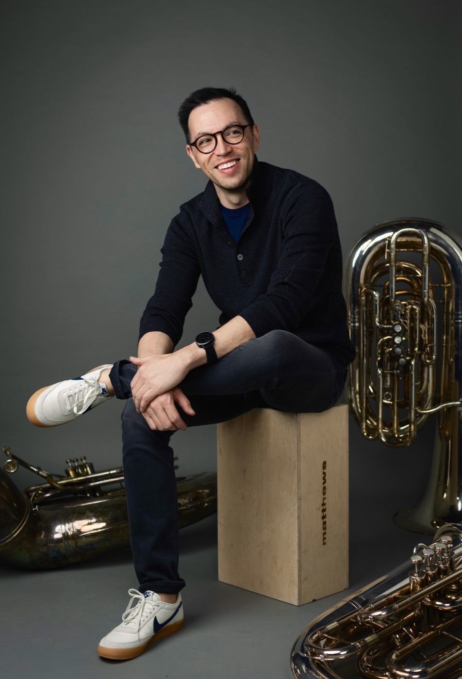 Neil Konouchi seated on a wooden box in a photo studio, surrounded by tubas